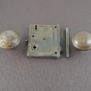 May include: An antique door lock set comprising two round knobs, a rectangular lock body, and a connecting rod. The metal components display a weathered, aged appearance with brown and grey tones, indicative of vintage hardware.
