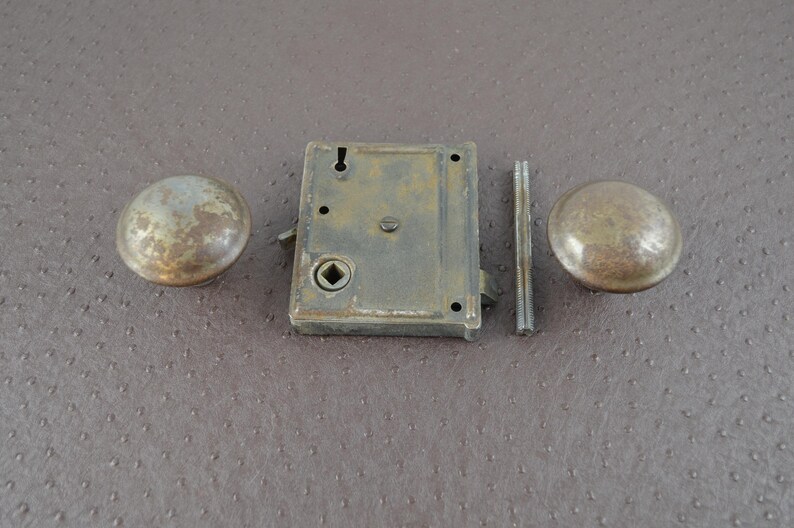 May include: An antique door lock set comprising two round knobs, a rectangular lock body, and a connecting rod. The metal components display a weathered, aged appearance with brown and grey tones, indicative of vintage hardware.