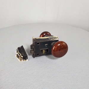 May include: A dark brown metal rim lock mechanism featuring two round, polished, wood-grain patterned knobs. The rectangular lock body has a brass-colored latch and a separate matching strike plate, both showing remnants of white paint along the edges.