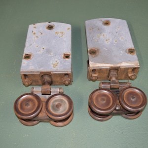 May include: Two vintage metal sliding door rollers with brown wheels. The rollers are attached to metal plates with screws and have a metal arm connecting the two wheels.