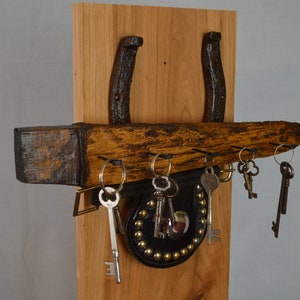 May include: A rustic key holder made from a wooden plank with two horseshoes attached to the back. The plank has several hooks for hanging keys. There is a small leather pouch with metal studs hanging from one of the hooks.