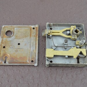 May include: A vintage door lock mechanism, in pieces. The lock comprises a rectangular metal housing with a keyhole, a separate cover plate, and internal brass-coloured levers and springs. The metal shows signs of age and wear, with some rust.