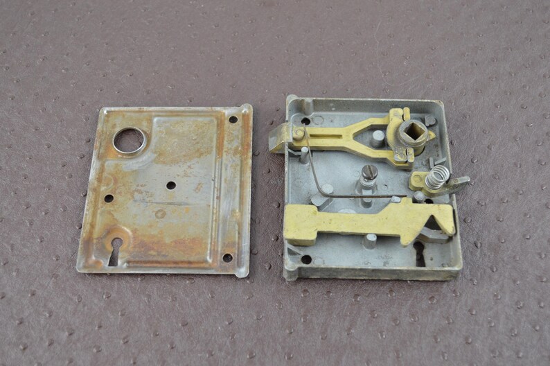 May include: A vintage door lock mechanism, in pieces. The lock comprises a rectangular metal housing with a keyhole, a separate cover plate, and internal brass-coloured levers and springs. The metal shows signs of age and wear, with some rust.