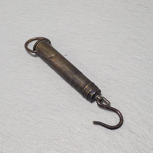 May include: A vintage brass spring scale with a cylindrical body, a top ring, and a curved hook at the bottom. The scale has a worn, antique appearance, suggesting age and use. The design is simple and functional.