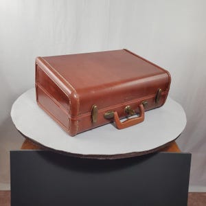 May include: A vintage brown suitcase with brass hardware and a brown handle. The suitcase has a rectangular shape with rounded corners and white stitching around the edges. The suitcase is sitting on a round, white surface.
