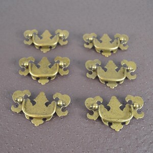 May include: Six antique brass cabinet pulls with ornate designs. Each pull has a small, rectangular opening for a screw.