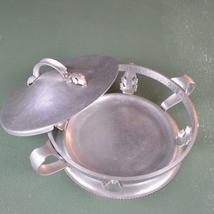 May include: A silver aluminum chafing dish with a lid and two handles. The dish has a decorative design around the rim.