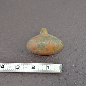 May include: A small, antique, copper-colored flask with a rounded body and a narrow neck. The flask has a weathered appearance with green oxidation. A measuring tape is next to the flask, showing inches.