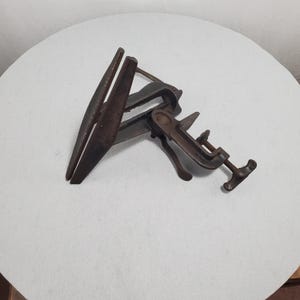 May include: An antique metal clamp with a dark, aged finish. The clamp features a screw mechanism and two angled jaws, suggesting it's designed for holding objects. The overall design indicates a vintage tool, likely used for woodworking or similar crafts.