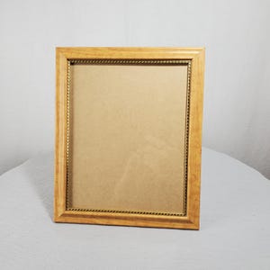 May include: A rectangular wooden picture frame with a light brown finish. The frame has a gold-colored rope-like detail around the inner edge. The frame is empty, with a tan backing visible inside.