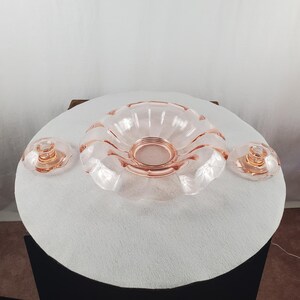 May include: A set of three pieces of pink depression glass, including one large scalloped bowl with a central circular base and two matching smaller candle holders, each featuring a fluted design and a smooth, translucent finish.