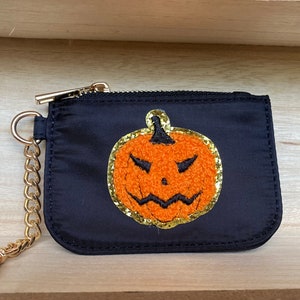 May include: A black zippered pouch with a gold chain and a gold embroidered pumpkin with a scary face.