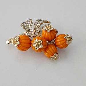 May include: A gold-toned brooch featuring a cluster of orange pumpkin-shaped beads with gold accents. The brooch includes a stem and leaves embellished with clear crystals, creating a festive autumn theme.