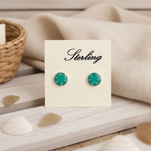 Puede incluir: Pendientes de botón de plata de ley con centros de color turquesa. Los pendientes se muestran en una tarjeta color crema con la palabra "Sterling". El fondo incluye una cesta tejida, conchas marinas y una superficie de madera.