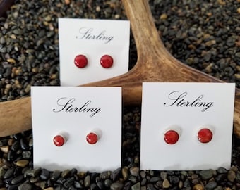 May include: Three pairs of red stud earrings on white cards with the word "Sterling" in cursive. The earrings are displayed on a bed of small, dark pebbles, with a wooden antler in the background.