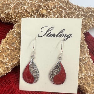 May include: A pair of sterling silver drop earrings featuring a teardrop shape. Each earring has a red stone inlay paired with a silver filigree cutout design. The earrings are attached to a white display card with the word Sterling printed.