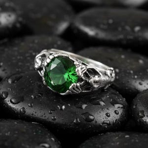 Puede incluir: Un anillo plateado con una gran gema verde esmeralda facetada. El anillo tiene un diseño ornamentado y detallado alrededor de la gema. El anillo está sobre un fondo de piedras negras mojadas.
