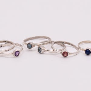Op de afbeelding: Zes zilveren ringen met verschillende gekleurde edelstenen. De ringen liggen op een witte ondergrond. De edelstenen zijn blauw, paars, rood en wit.