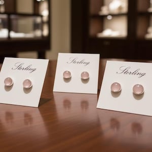 Può includere: Tre paia di orecchini a bottone in argento sterling con pietre preziose rosa e rotonde esposti su cartoncini bianchi. La parola "Sterling" è scritta in elegante corsivo su ogni cartoncino. Gli orecchini sono su una superficie di legno scuro.