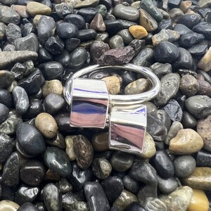 May include: A silver ring with two square-shaped bands that are connected by a thin band. The ring is sitting on a bed of small, dark grey stones.
