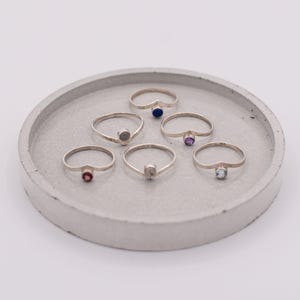 May include: Six silver rings with different coloured gemstones are arranged on a grey, circular tray. The gemstones include red, grey, blue, purple, and clear stones. The rings are displayed against a white background.