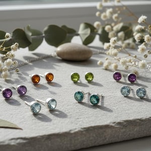 May include: A collection of gemstone stud earrings in various colors, including orange, green, purple, and blue. The earrings are set in silver and arranged on a gray surface, with floral accents in the background.