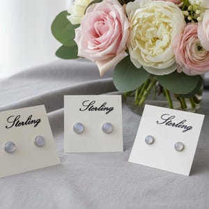 May include: Three pairs of sterling silver stud earrings with a light blue iridescent finish, displayed on white cards with the word "Sterling" in elegant script. A bouquet of pink and white roses and greenery is in the background.