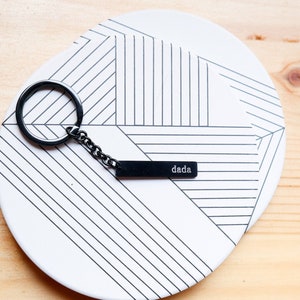 May include: A black metal keychain with a rectangular tag that says "dada". The keychain is on a white plate with a black and white geometric pattern.