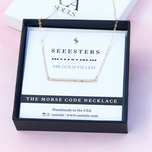 以下が含まれることがあります： モールス信号で「SEEESTERS」と書かれたバーペンダントが付いたゴールドネックレス。ネックレスは、黒色の箱に入っていて、白いラベルには「14K GOLD FILLED」と「THE MORSE CODE NECKLACE」と書かれています。ラベルには「Handmade in the USA」とウェブサイトのアドレス「casouls | www.casouls.com」も記載されています。
