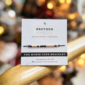 Może przedstawiać: Bransoletka w kolorze czarnym i złotym z napisem "BROTHER", "14K GOLD FILLED - GLASS BEADS" i "THE MORSE CODE BRACELET" na białej kartce. Na kartce znajduje się również tekst "Handmade in the USA" i "casouls | www.casouls.com".