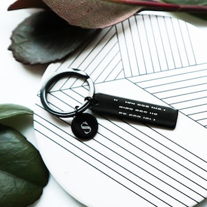 Può includere: Portachiavi in metallo nero con un piccolo ciondolo rotondo inciso con la lettera "S" e un'etichetta rettangolare con lettere in braille.