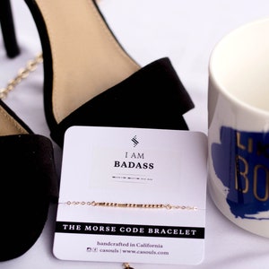 May include: A white card with black text that reads "I am badass" and "The Morse Code Bracelet". The card is on a white surface with a pair of black heels and a white mug with blue and gold text that reads "Like a Boss".
