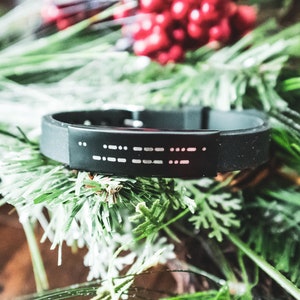 Puede incluir: Una pulsera de silicona negra con una placa plateada que presenta un mensaje en braille.