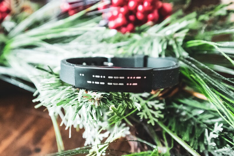 Puede incluir: Una pulsera de silicona negra con una placa plateada que presenta un mensaje en braille.