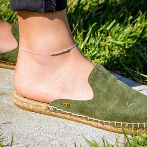 May include: A silver anklet with small beads on a chain. The anklet is worn on a person's ankle with green suede shoes.