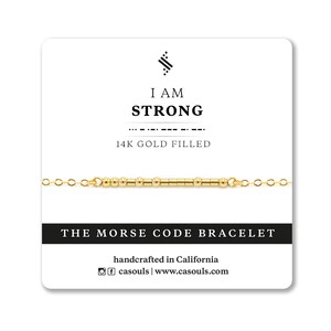 Può includere: Braccialetto dorato con una barra di perline che compongono la scritta "I AM STRONG" in codice Morse. Il braccialetto è su uno sfondo bianco con il testo "14K GOLD FILLED", "THE MORSE CODE BRACELET", "handcrafted in California", "casouls | www.casouls.com".