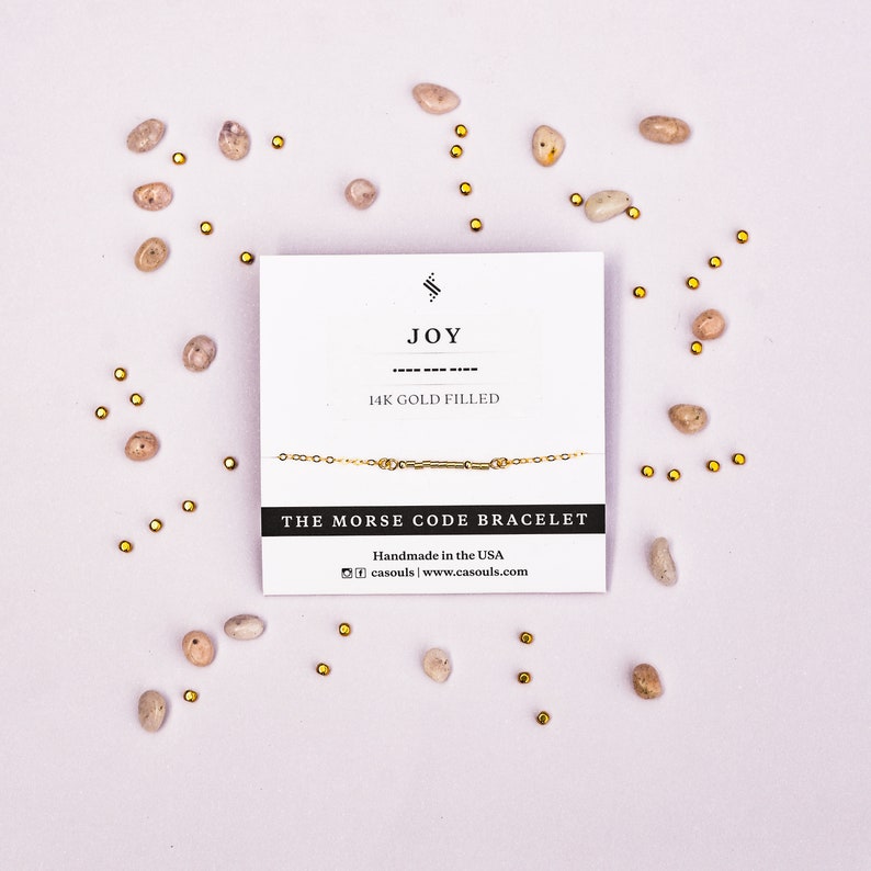 May include: A gold-filled bracelet with the word "JOY" spelled out in Morse code. The bracelet is on a white card with the text "14K GOLD FILLED" and "THE MORSE CODE BRACELET". The card also includes the text "Handmade in the USA" and the website address "www.casouls.com".