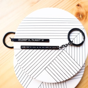 May include: A black metal keychain with a silver bar engraved with the coordinates "39.6480° N, 79.9697° W".