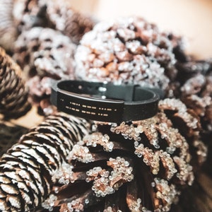 Puede incluir: Una pulsera de silicona negra con una banda plateada grabada con puntos braille. La pulsera est&aacute; apoyada sobre un lecho de pi&ntilde;as marrones.