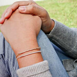May include: A person wearing a gray sweater and blue jeans with two bracelets on their wrist. The bracelets are made of small silver and gold beads.