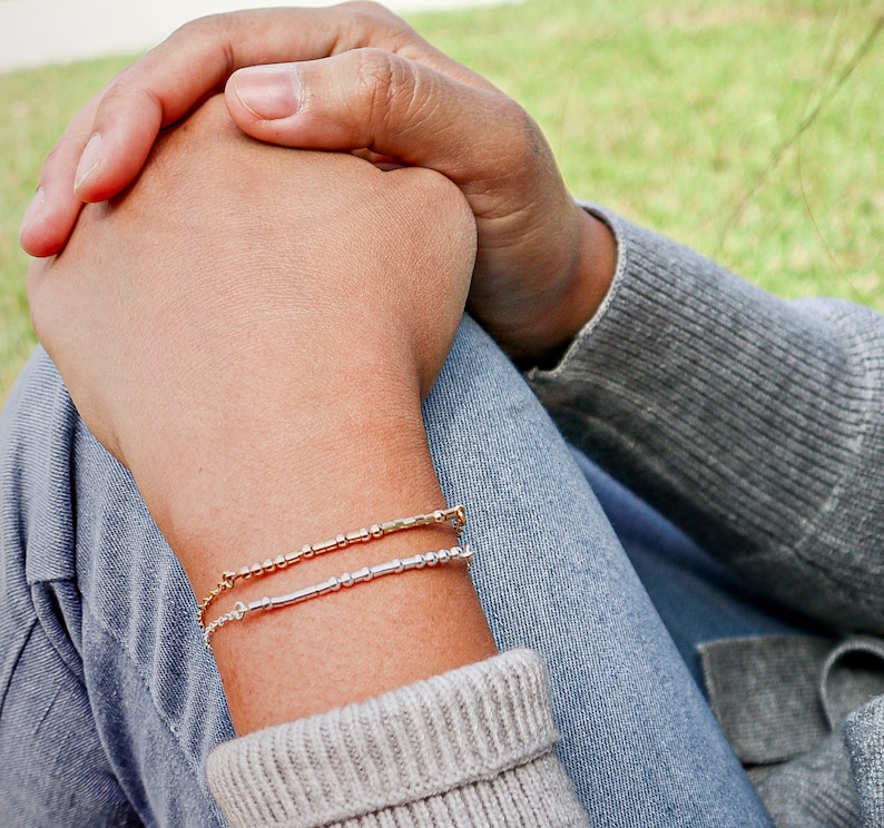May include: A person wearing a gray sweater and blue jeans with two bracelets on their wrist. The bracelets are made of small silver and gold beads.