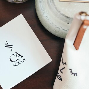 May include: A white box with a black logo that says "CA SOULS" and a stylized design of three lines with dots on each end. The box is on a dark brown wooden surface.