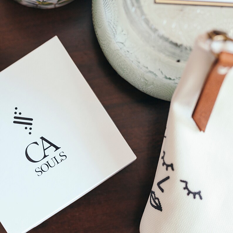 May include: A white box with a black logo that says "CA SOULS" and a stylized design of three lines with dots on each end. The box is on a dark brown wooden surface.