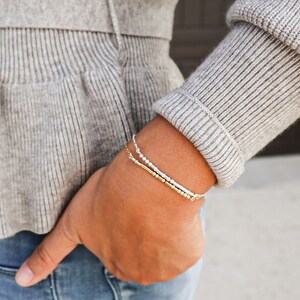 May include: A close-up of a person's wrist wearing two bracelets. The top bracelet is made of silver beads and the bottom bracelet is made of gold beads.