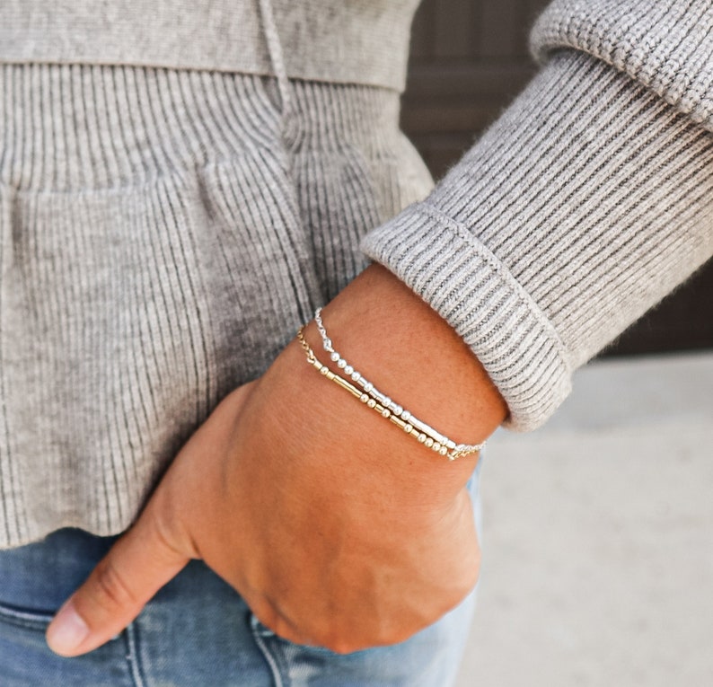 May include: A close-up of a person's wrist wearing two bracelets. The top bracelet is made of silver beads and the bottom bracelet is made of gold beads.