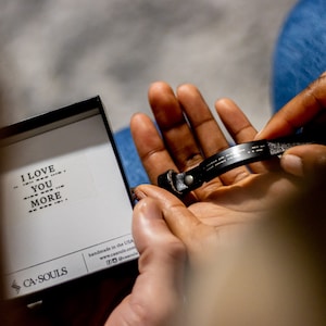 Puede incluir: Un brazalete de cuero negro con las palabras "I love you more" escritas en braille. El brazalete est&aacute; en una peque&ntilde;a caja blanca con las palabras "CA SOULS" y "hecho a mano en los EE. UU." impresas en ella.