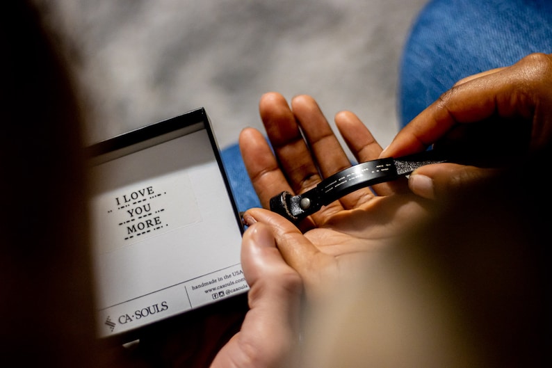 Puede incluir: Un brazalete de cuero negro con las palabras "I love you more" escritas en braille. El brazalete est&aacute; en una peque&ntilde;a caja blanca con las palabras "CA SOULS" y "hecho a mano en los EE. UU." impresas en ella.