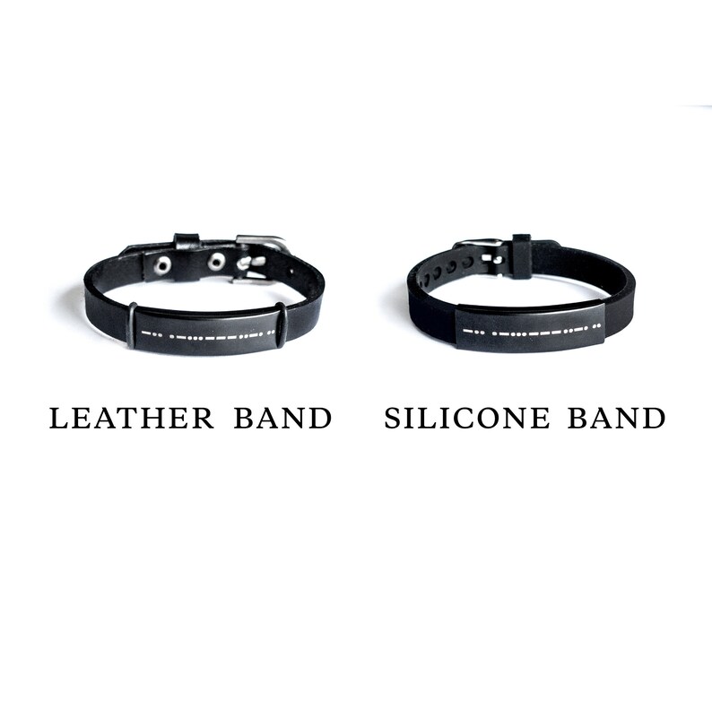 Puede incluir: Dos pulseras negras con una hebilla plateada y una placa plateada con letras en braille. La pulsera de la izquierda es una banda de cuero y la pulsera de la derecha es una banda de silicona.