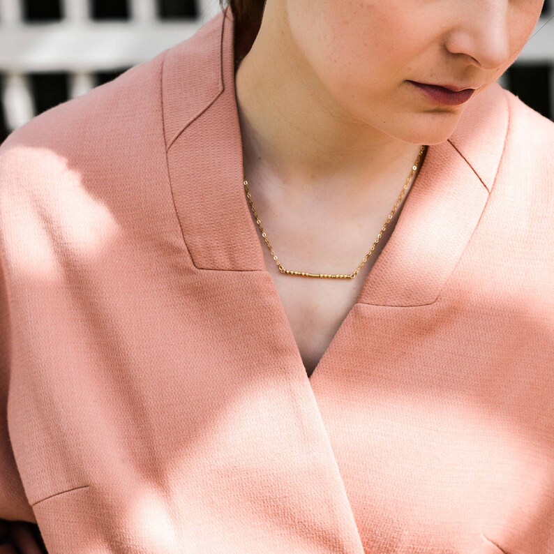 May include: A close-up of a person wearing a light pink blazer with a gold chain necklace featuring a small bar pendant.