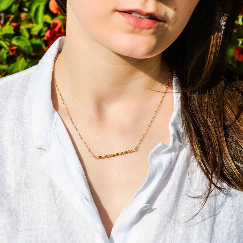 May include: A gold necklace with a delicate chain and a thin bar pendant. The necklace is worn by a person with long brown hair.